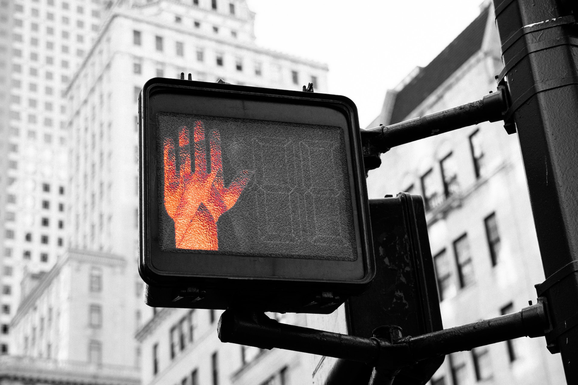 Description: the orange hand of a 'do not walk' sign.  The gray buildings in the background are slightly out of focus. Photo by Kai Pilger on Unsplash