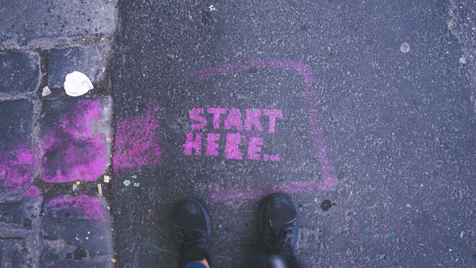 Description: an overhead photo of a street with the words 'start here' written in hot-pink paint/chalk.  A person's running shoes are seen at the bottom of the frame. Photo by Gia Oris on Unsplash.